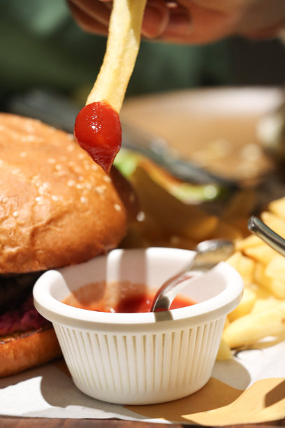 特写照片中，一个无法辨认的人把酥脆、金黄的薯条放在番茄酱的小模子里，奶酪汉堡放在芝麻汉堡包里，一堆炸土豆片放在前景的焦点上图片下载