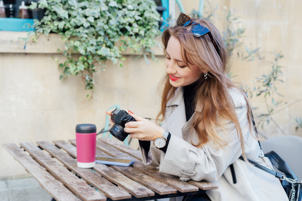 快乐微笑的女性旅行者在城市散步后，在咖啡馆露台上拿着可重复使用的杯子里的咖啡休息，看着数码相机上的照片。旅游博主、摄影师、网红捕捉这一刻。图片下载