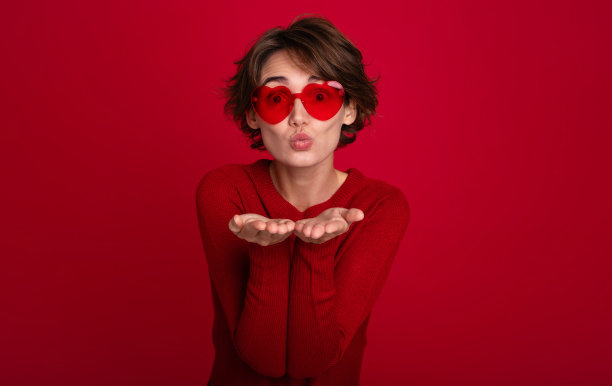 近距离的照片，迷人的时髦女人戴着心形的眼镜，她在红色明亮的墙壁上孤独地飞吻。女孩心情好，有爱的情绪图片下载