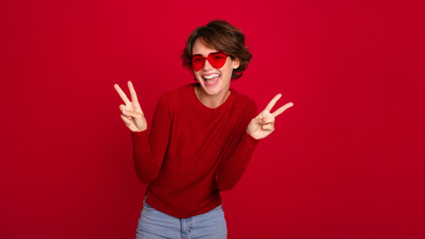 照片中，一位戴着心形眼镜的时髦女性微笑着，兴奋地笑着，胜利的标志被隔离在明亮的红色墙壁上。女孩心情好，有爱的情绪图片下载