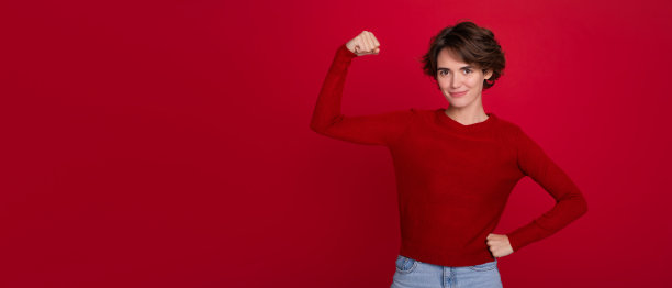 横幅广告照片上，一个自信的现代女性，留着时髦的短发，穿着红色毛衣，她的二头肌被红色背景孤立起来:女权，女权主义。图片下载