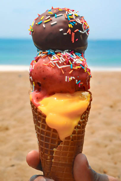 特写照片中，无法辨认的人拿着华夫蛋筒、三勺巧克力、芒果和草莓冰淇淋、五彩糖粒、晴朗的蓝天和沙滩背景，重点放在前景上图片下载