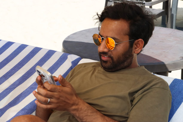 特写照片:印度男子戴着黄色镜片的镜面太阳镜，斜倚在太阳躺椅上，铺着合身的蓝白条纹毛巾，正在使用手机，这是前景的焦点图片下载