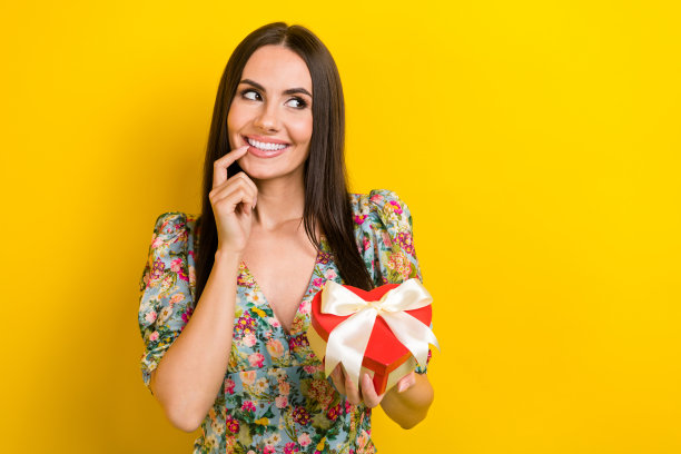 照片上的漂亮女孩留着长发，穿着印花衬衫，抱着心盒子，在黄色的背景上看起来很空旷图片下载