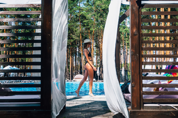 漂亮的年轻女子穿着黑色泳衣，戴着帽子，戴着太阳镜，沿着室外游泳池的边缘走着。夏日、度假和生活方式的概念图片下载