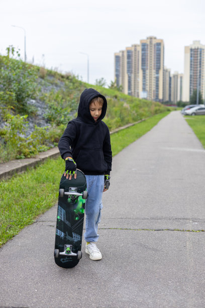 一个时髦的男孩在城市街道上用脚拿着滑板。垂直的。一个孩子在城市里玩滑板的漂亮照片。一个孩子骑滑板的概念。图片下载