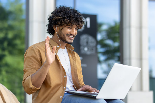 一个年轻的穆斯林男子坐在外面的长椅上，戴着耳机，对着摄像机挥手，正在用笔记本电脑进行视频通话图片下载