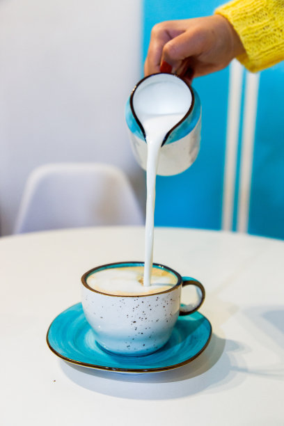 这张美丽的照片是在咖啡店里，白色的桌子上放着一个绿松石色的茶托，把陶瓷水罐里的卡布奇诺咖啡倒入杯子里，溅起水来，溢出来图片下载