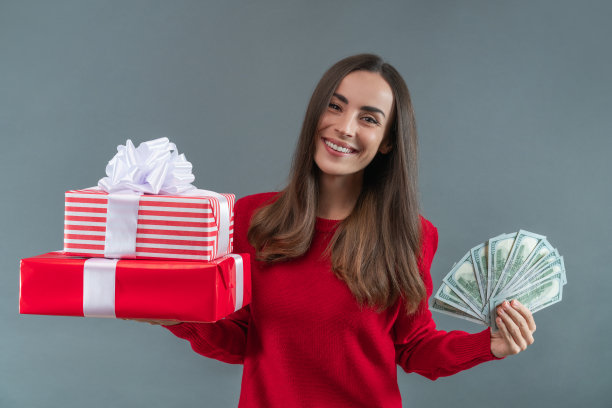 一个微笑的快乐的年轻女子的肖像，手里拿着美元钞票和一堆礼盒，孤立在灰色的背景上图片下载