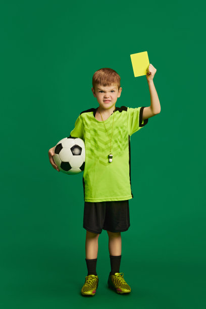 男孩打扮成足球裁判，拿着球和黄牌，愤怒的表情在充满活力的绿色背景下。初级裁判。图片下载