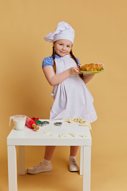 戴着厨师帽的小女孩，端着新鲜出炉的美味羊角面包，在米色工作室背景下梦想着烹饪的未来。图片下载