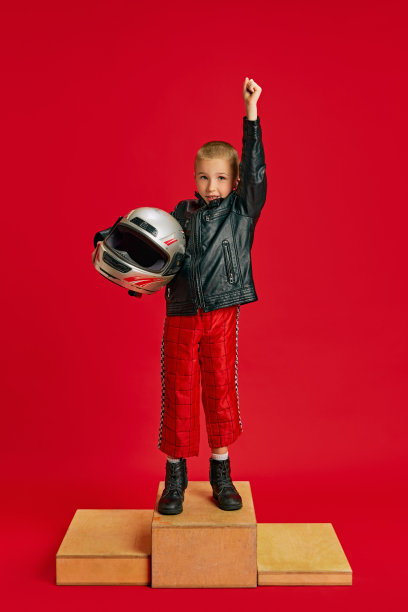孩子，小男孩穿着赛车服站在木制舞台上庆祝胜利，在充满活力的红色背景下想象比赛。图片下载