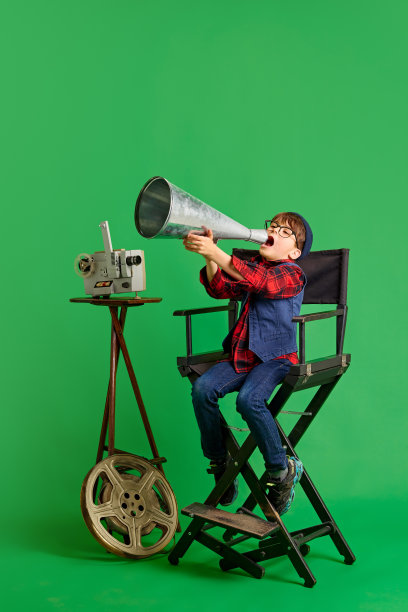 一个打扮成导演的男孩坐在导演的椅子上，在绿色的背景下对着老式的扩音器大声喊叫。图片下载