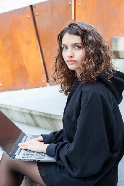 照片中，一位美丽的卷发少女在城市的街道上使用笔记本电脑。图片下载