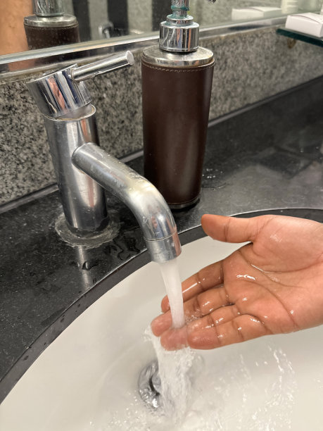 特写照片中，一个无法辨认的人正在测试水龙头的水温，从镀铬的单杆龙头，酒店浴室的水槽，镜子前的黑色花岗岩洗手台上的皂液器，大理石墙砖，重点放在前景上图片下载