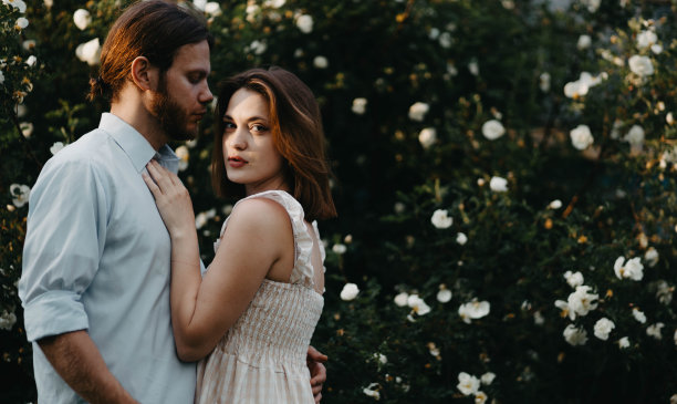 美丽的夫妇穿着复古的衣服拥抱在一棵白玫瑰树在夏天的村庄。图片下载