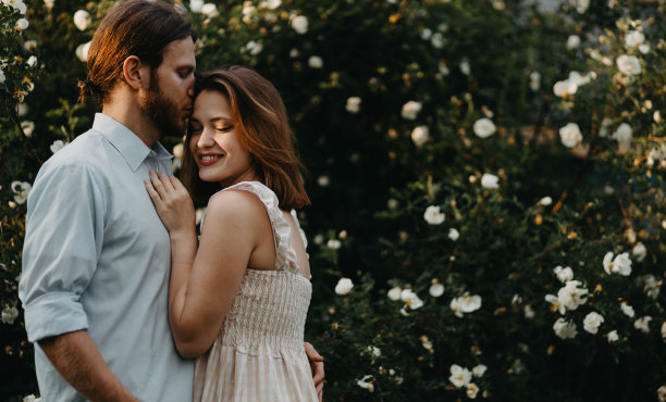 美丽的夫妇穿着复古的衣服拥抱在一棵白玫瑰树在夏天的村庄。图片下载