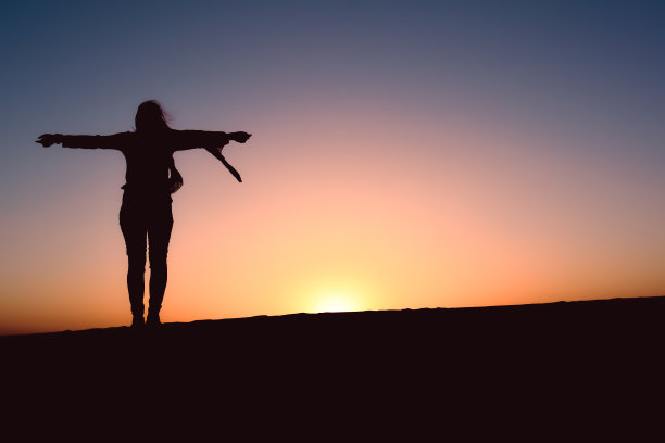 站着的女人独自一人的身影，双手摊开双臂在沙丘上欣赏日落的观点，享受户外的自由。概念自由和无忧无虑的生活方式旅游概念图片下载