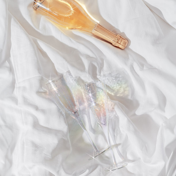 彩虹色闪闪发光的香槟酒杯和白色闪闪发光的酒瓶放在床上，白色的毯子为背景。生活美学照片，明星滤镜。浪漫的聚会，浪漫的节日理念。图片下载