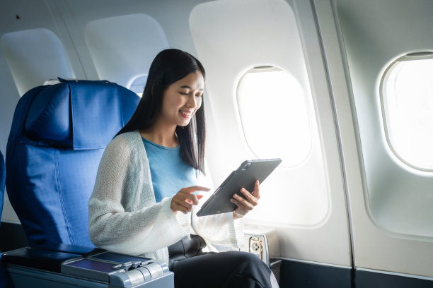 一位年轻的亚洲女乘客在飞行过程中坐在窗口，拿着登机牌，头顶上放着随身行李，系着安全带，享受着旅程。图片下载