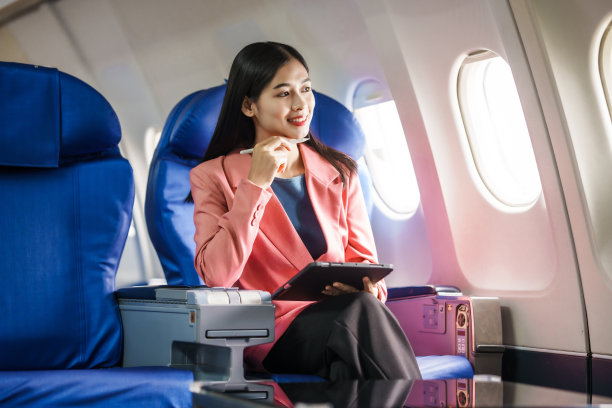 一位年轻的亚洲女乘客在飞行过程中坐在窗口，拿着登机牌，头顶上放着随身行李，系着安全带，享受着旅程。图片下载