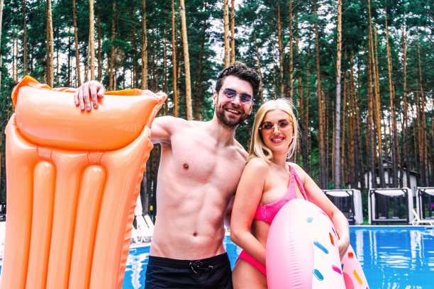 一对戴着墨镜微笑的年轻夫妇在室外游泳池边摆姿势，手里拿着充气床垫和戒指。暑假，积极休闲，生活理念图片下载