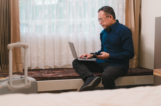 一个男人正在长凳上工作，手里拿着一台笔记本电脑和一部手机。他在看笔记本电脑的屏幕图片下载