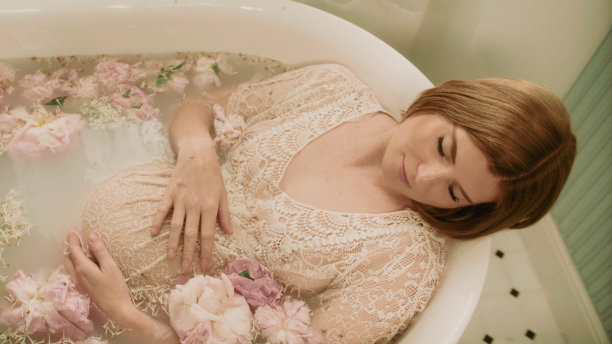 一个红头发的孕妇在一个装满粉红色花朵的浴缸里洗澡——牡丹和牛奶。图片下载