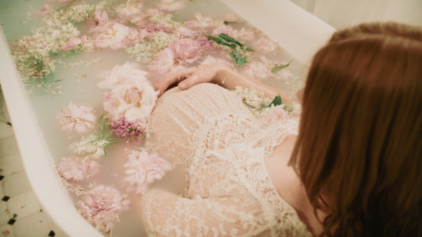 一个红头发的孕妇在一个装满粉红色花朵的浴缸里洗澡——牡丹和牛奶。图片下载