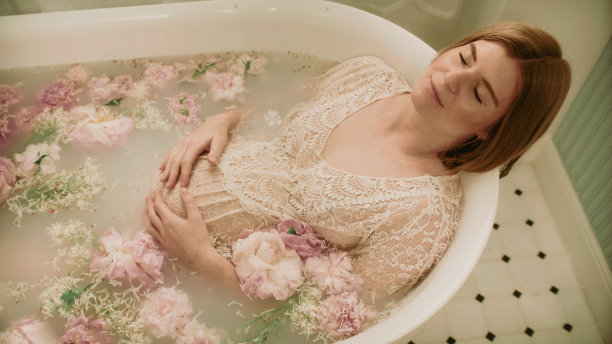 一个红头发的孕妇在一个装满粉红色花朵的浴缸里洗澡——牡丹和牛奶。图片下载