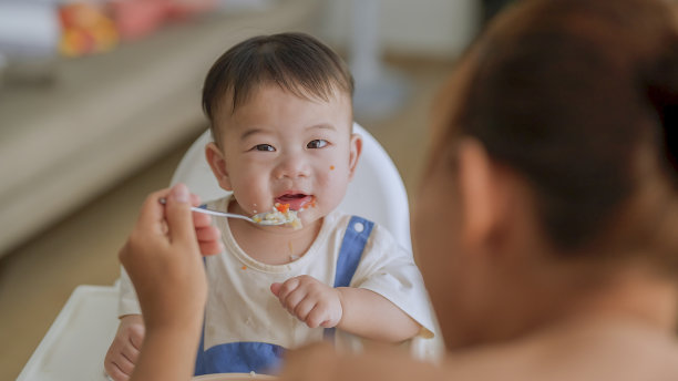 亚洲妈妈在家用高脚椅上用勺子喂凌乱的女婴的照片宝宝里程碑，成长和发展的概念图片下载