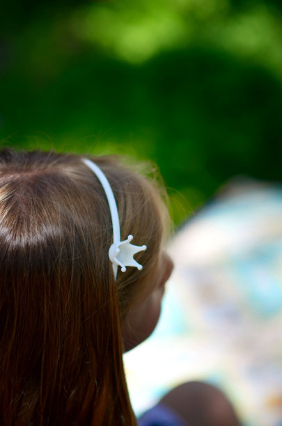 这是一张特写照片，拍摄了一个戴着白色王冠的孩子的后脑勺，背景是模糊的自然背景。图片下载