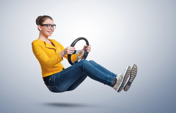 戴着眼镜的年轻快乐微笑的女孩开着一辆车，手里拿着方向盘，背景是浅蓝色的。汽车的概念图片下载