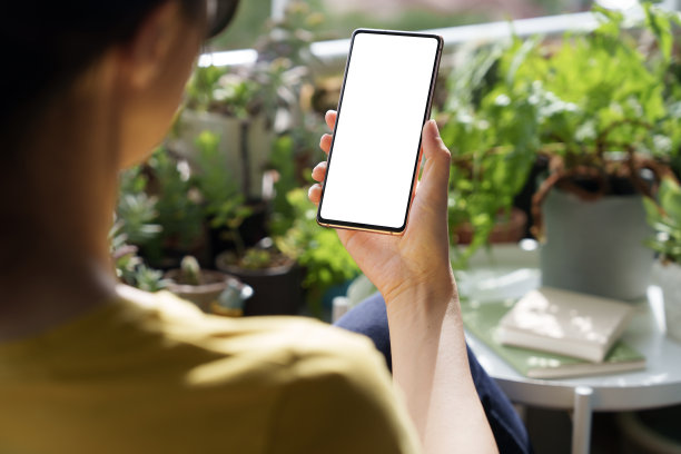 在这张裁剪过的图片中，一名亚洲女子放松地坐在花园后院的野餐椅上，使用着一部空白屏幕的智能手机。图片下载