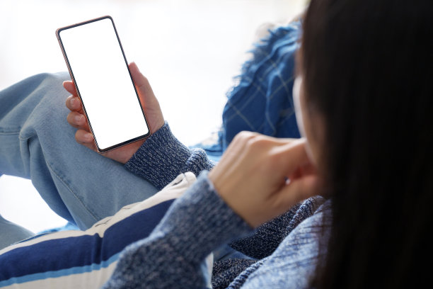 在这张裁剪过的图片中，一名亚洲女性放松地坐在沙发上，使用着一部空白屏幕的智能手机。图片下载
