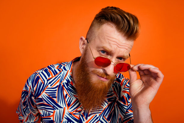 照片忧郁的家伙与红胡子时髦的发型，穿着时尚的衬衫触摸太阳镜凝视孤立的充满活力的橙色背景图片下载