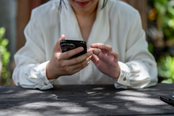 这是一张裁剪过的照片，一个晴朗的日子里，一名女子坐在花园的一张木桌旁使用智能手机。图片下载