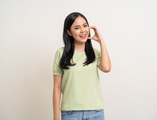 漂亮的年轻美丽的亚洲女人站着微笑，在孤立的白色背景上显示迷你心脏标志。可爱的拉丁女性穿着绿色衬衫感到惊讶和快乐。图片下载