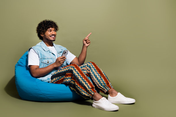 年轻人的全身照片坐在豆袋装置点看空空间穿着时尚的牛仔衣服孤立在卡其色背景图片下载