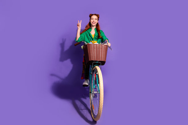 好心情的女孩穿着衬衫印花裤子骑自行车的全身照片显示v字符号孤立的紫色背景图片下载