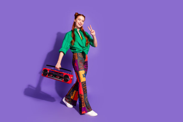全尺寸照片，乐观的女人穿着绿色衬衫印花裤子，拿着音箱，在紫色的背景上显示v字图片下载