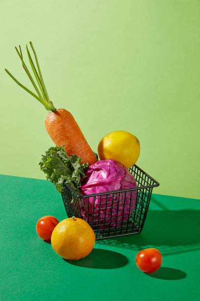 以蔬菜和水果为主题的绿色的色调照片，从对角线角度拍摄。在绿色的桌子中间有一个黑色的网状篮子，里面放着新鲜的蔬菜，旁边还有一些西红柿和一个橙子图片下载