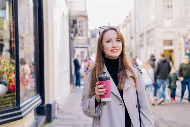 优雅的年轻微笑女子穿着风衣，一边走在城市街道上，一边用可重复使用的杯子喝热咖啡。时令流行风格趋势。可持续的日常生活方式。外带咖啡图片下载