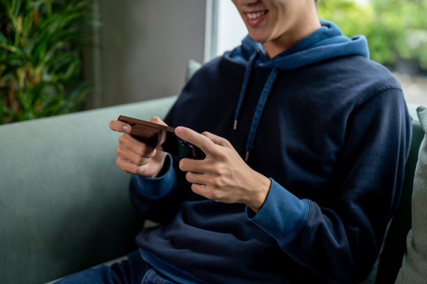 一张裁剪过的照片上，一位年轻、开朗的亚洲男子正坐在家里的沙发上玩手机游戏。图片下载