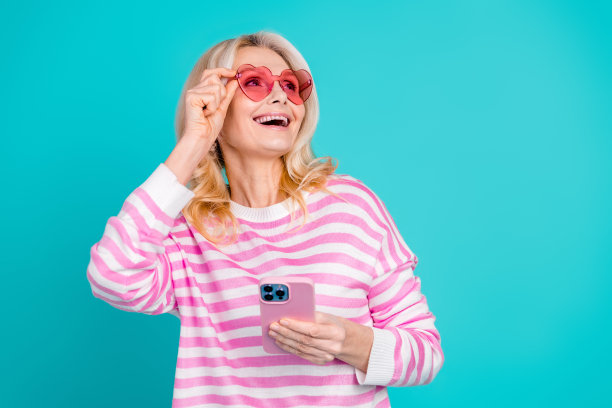 照片中，梦幻般的资深女性穿着条纹运动衫，手持智能手机，触摸太阳镜，在蓝色背景下显得空荡荡图片下载