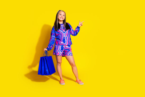 可爱的女人穿着印花衬衫的全尺寸照片，拿着购物袋，表明在黄色背景上的空白空间图片下载