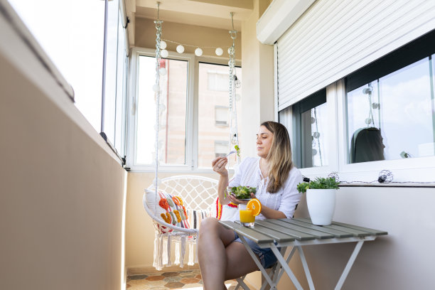 一个女人在阳台上享受着清晨美丽的阳光。她喜欢吃健康的蔬菜沙拉。橙汁也在桌子上。穿便服的女子，穿白衬衫。库存图片图片下载