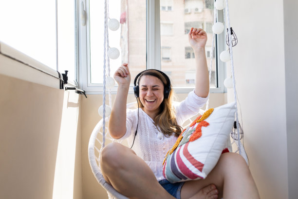 这位女士早餐后坐在露台上，戴着耳机听音乐。她玩得很开心。美丽的女人跳舞和微笑，早晨的城市氛围。库存图片图片下载