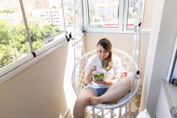 一个女人在阳台上享受着清晨美丽的阳光。她喜欢吃健康的蔬菜沙拉。穿便服的女子，穿白衬衫。库存图片图片下载