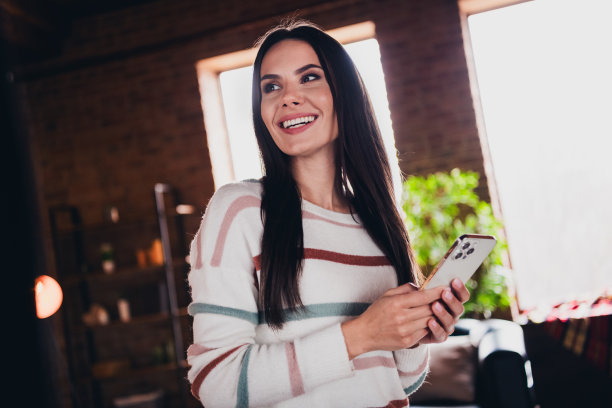 可爱的年轻女子拿着电话购物的照片，看着空旷的空间，穿着棕色暖色调的条纹装在家里客厅里图片下载